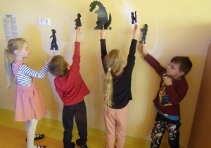 Czworo dzieci trzyma w ręku sylwety ludzi i zwierząt na które święcą latarkami tworząc cień na ścianie.